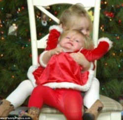 This little girl is determined to not sit still while her older sister clings to her for dear life as she flails around  in front of a Christmas tree while the photographer snaps away regardless